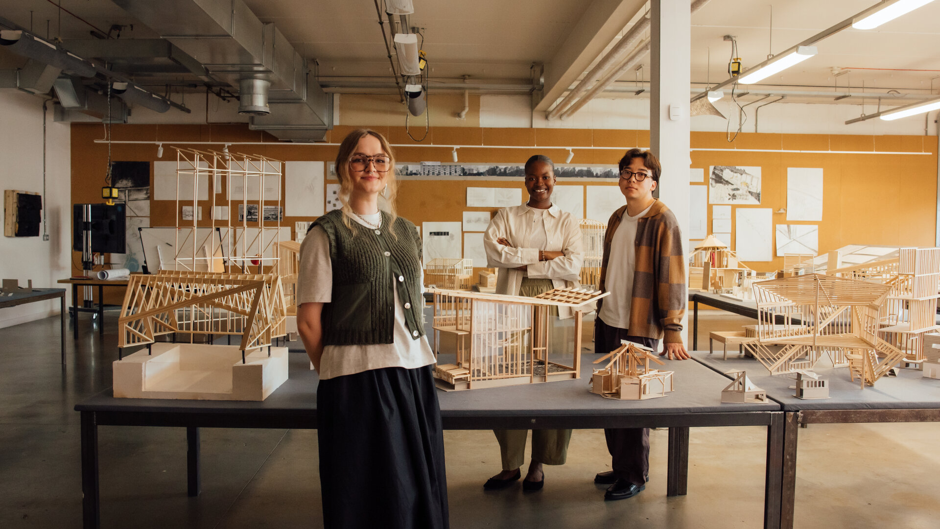 Three students stand smiling among tables of intricate wooden architectural models in a design studio, with a pinboard of drawings and workshop equipment in the background.